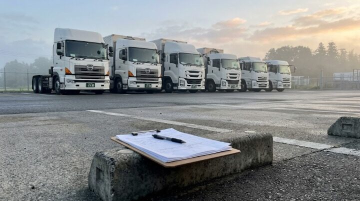 早朝のトラックヤードに並ぶ大型トラックと、手前の車止めに置かれた申請書類とペン